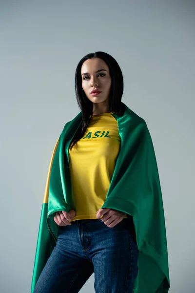 Schöne weibliche Fußballfan in brasilianische Flagge eingehüllt isoliert auf grau — Stockfoto