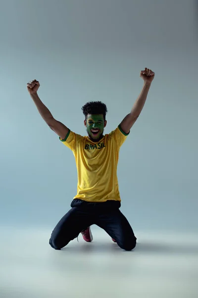 Excited african american football fan with face painted as brazilian flag kneeling on grey — Stock Photo