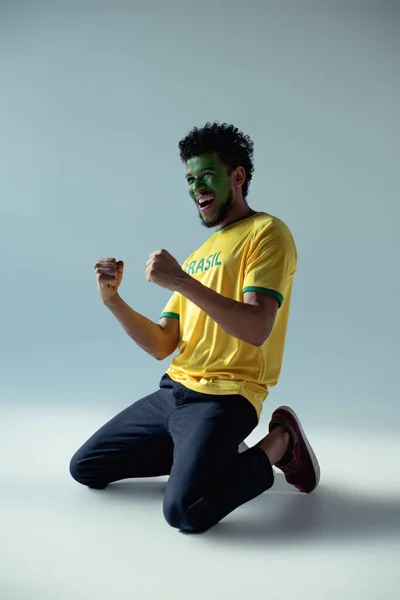 Émotionnel afro-américain fan de football avec le visage peint comme drapeau brésilien agenouillé sur gris — Photo de stock