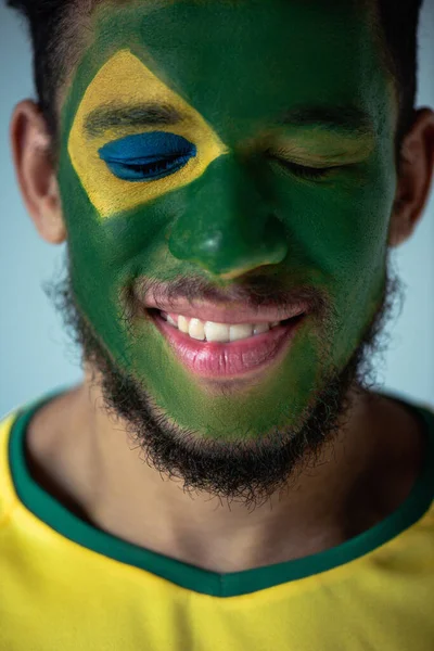 Усміхнений афроамериканський футбольний фанат із закритими очима та обличчям, намальованими як бразильський прапор, ізольований на сірому — стокове фото