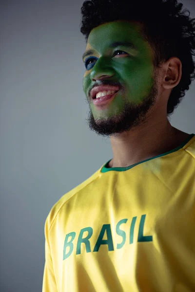 Позитивный африканский американский футбольный фанат с лицом, окрашенным как бразильский флаг, изолированный на сером — стоковое фото