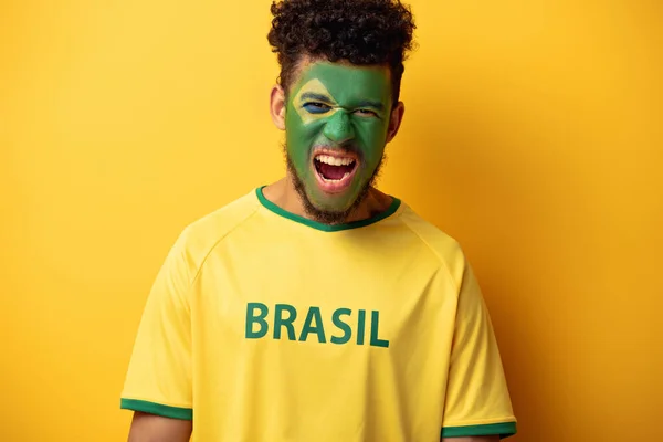 Tifoso afroamericano di calcio con faccia dipinta in t-shirt con cartello brasiliano che urla sul giallo — Foto stock