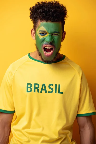 Emotional african american football fan with painted face in t-shirt with brazil sign shouting on yellow — Stock Photo