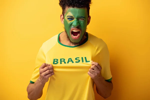 Aggressive african american football fan with painted face in t-shirt with brazil sign shouting on yellow — Stock Photo