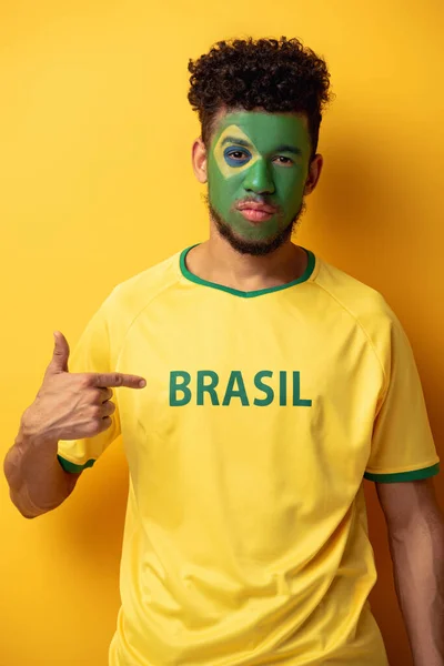Serio tifoso afroamericano di calcio con volto dipinto che punta a t-shirt con segno brasiliano su giallo — Foto stock