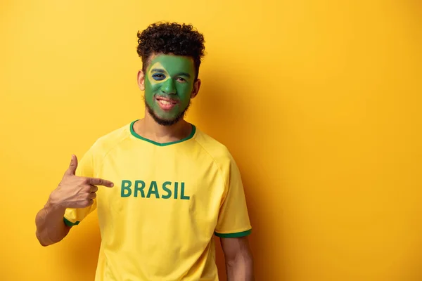 Lächelnder afrikanisch-amerikanischer Fußballfan mit aufgemaltem Gesicht, der auf ein T-Shirt mit brasilianischem Schriftzug in Gelb zeigt — Stockfoto