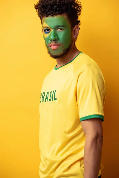 Handsome african american football fan with face painted as brazilian flag on yellow — Stock Photo