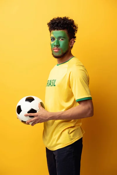 Bel tifoso afroamericano di calcio con la faccia dipinta come bandiera brasiliana che tiene la palla sul giallo — Foto stock