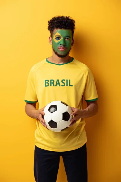 African american football fan with face painted as brazilian flag holding ball on yellow — Stock Photo