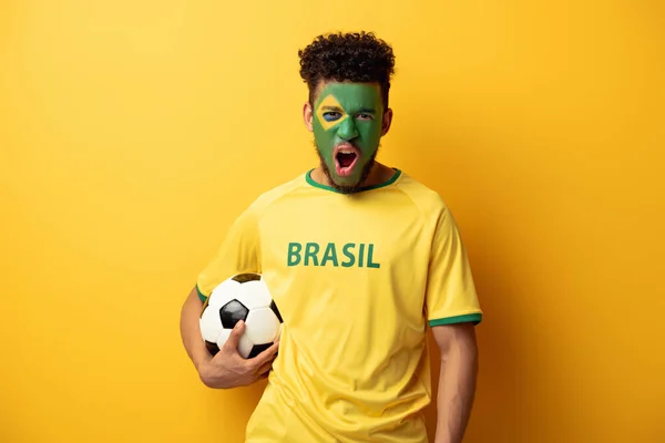 Emotional african american football fan with face painted as brazilian flag yelling and holding ball on yellow — Stock Photo