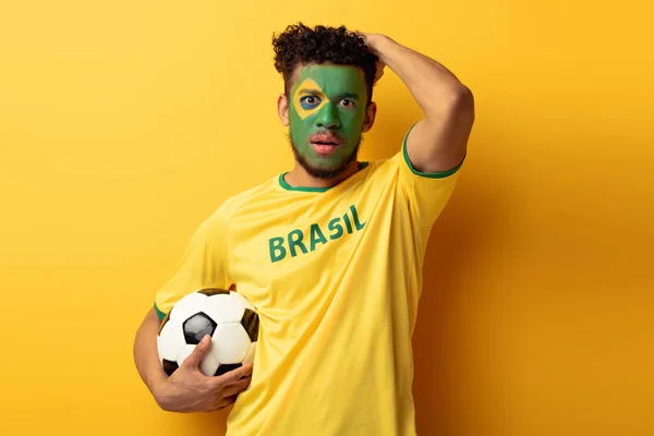 Шокований афроамериканський футбольний фанат з обличчям намальований як бразильський прапор, що тримає м'яч на жовтому — стокове фото