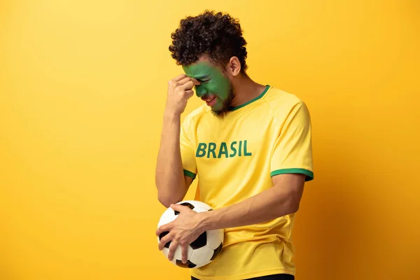 Confuso fã de futebol americano africano com rosto pintado como bandeira brasileira segurando bola no amarelo — Fotografia de Stock