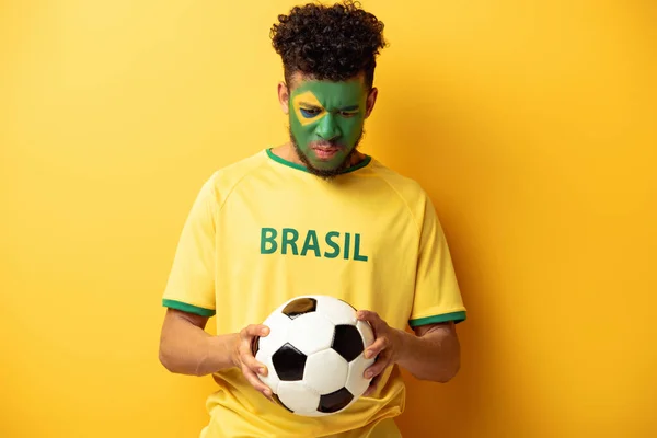 Sad african american football fan with face painted as brazilian flag holding ball on yellow — Stock Photo