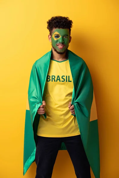 Felice tifoso afroamericano di calcio con volto dipinto avvolto in bandiera brasiliana su giallo — Foto stock