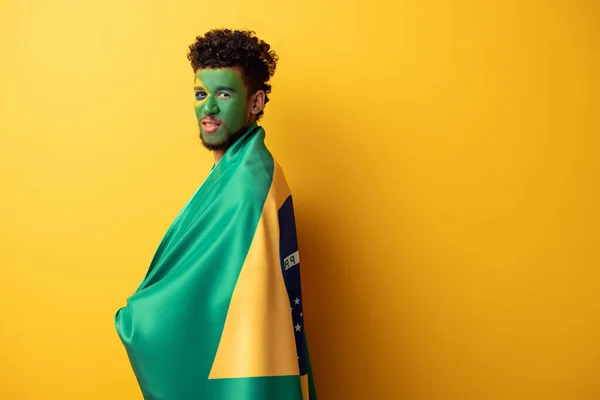 Apuesto fanático del fútbol afroamericano con la cara pintada envuelta en bandera brasileña en amarillo - foto de stock