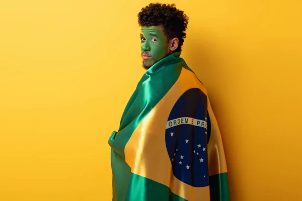 Fan de football afro-américain avec visage peint enveloppé dans le drapeau brésilien sur jaune — Photo de stock