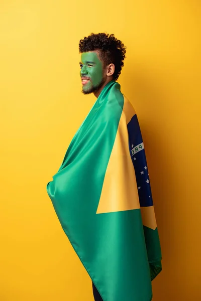 Abanico de fútbol americano africano sonriente con la cara pintada envuelta en bandera brasileña en amarillo - foto de stock