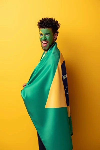 Appassionato di calcio afroamericano eccitato con volto dipinto avvolto in bandiera brasiliana su giallo — Foto stock