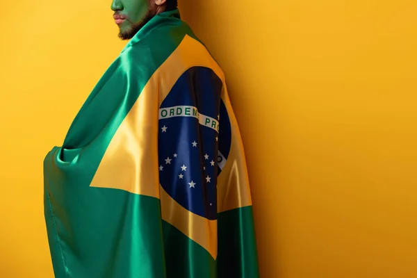 Cropped view of football fan with painted face wrapped in brazilian flag on yellow — Stock Photo