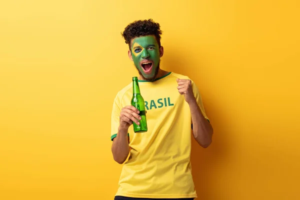 Torcedor de futebol americano africano alegre com rosto pintado como bandeira brasileira segurando garrafa de cerveja no amarelo — Fotografia de Stock