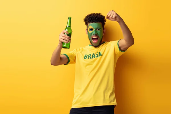 Fã de futebol americano africano animado com rosto pintado como bandeira brasileira segurando garrafa de cerveja no amarelo — Fotografia de Stock