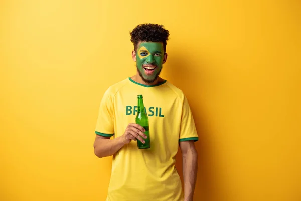 Fröhlicher afrikanisch-amerikanischer Fußballfan mit einem als brasilianische Flagge geschminkten Gesicht, das eine Flasche Bier auf gelb hält — Stockfoto