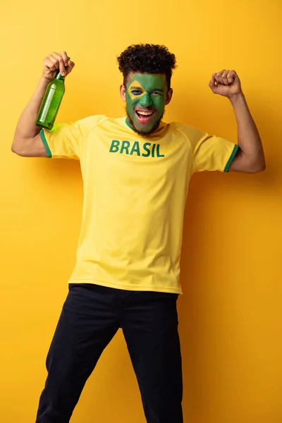 Aufgeregter afrikanisch-amerikanischer Fußballfan mit einem als brasilianische Flagge geschminkten Gesicht, das eine Flasche Bier auf gelb hält — Stockfoto