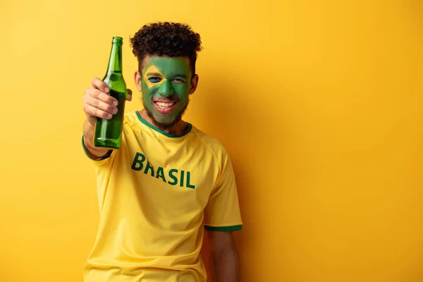 Smiling african american football fan with face painted as brazilian flag holding bottle of beer on yellow — Stock Photo