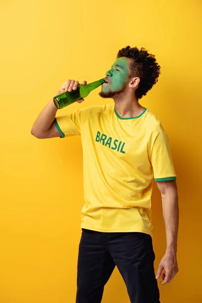 African american football fan with face painted as brazilian flag drinking beer on yellow — Stock Photo