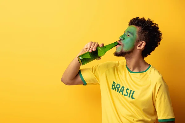 Fã de futebol americano africano feliz com rosto pintado como bandeira brasileira bebendo cerveja em amarelo — Fotografia de Stock