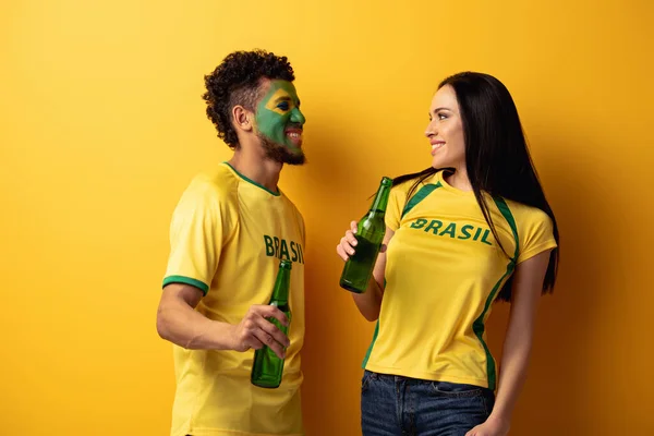 Male african american football fan with painted face and smiling woman holding bottles of beer on yellow — Stock Photo