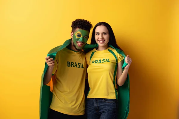 Abanico de fútbol americano africano masculino con la cara pintada y la niña sonriente sosteniendo bandera brasileña en amarillo - foto de stock