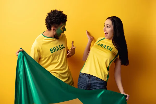 Tifoso di calcio afroamericano maschile con viso dipinto e ragazza felice gesticolando e tenendo bandiera brasiliana su giallo — Foto stock