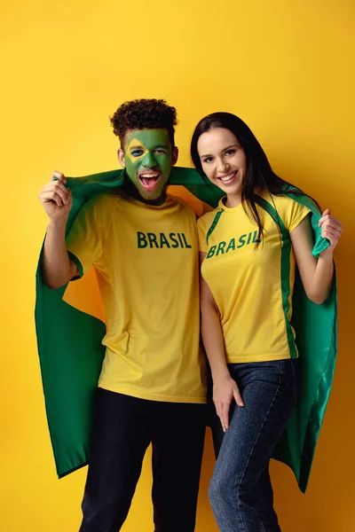 Apuesto hombre africano americano ventilador de fútbol con la cara pintada y feliz chica sosteniendo bandera brasileña en amarillo - foto de stock