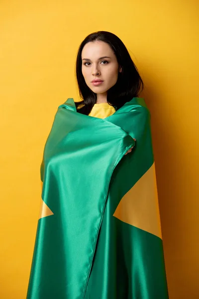 Attractive female football fan wrapped in brazilian flag on yellow — Stock Photo