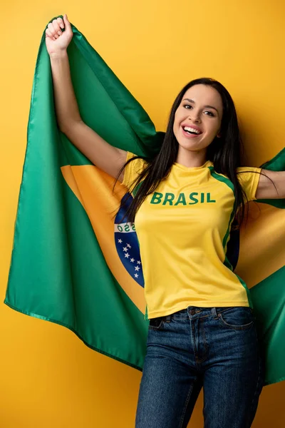 Atractiva y alegre hincha de fútbol femenino sosteniendo bandera brasileña en amarillo - foto de stock