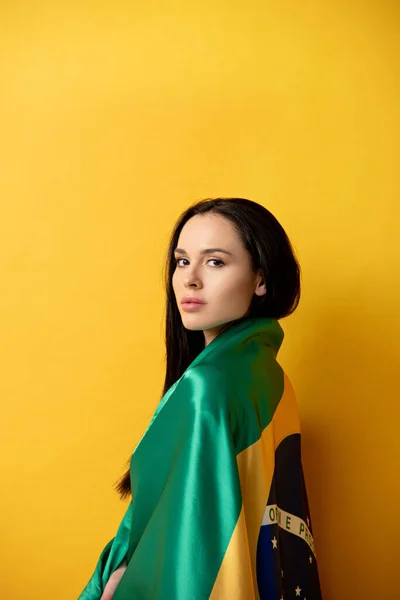 Attractive female football fan wrapped in brazilian flag on yellow — Stock Photo