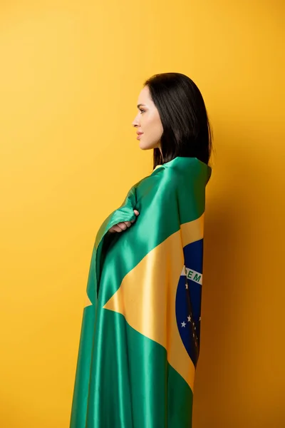 Smiling female football fan wrapped in brazilian flag on yellow — Stock Photo