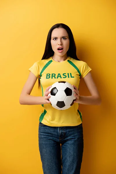 Шокований фанат жіночого футболу тримає м'яч на жовтому — стокове фото