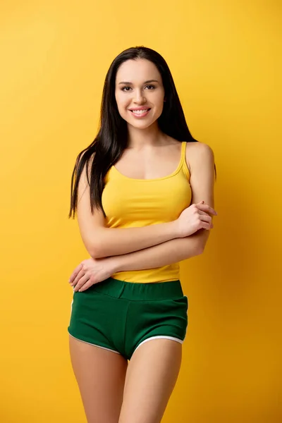 Приваблива усміхнена дівчина в зелених шортах, що стоїть з схрещеними руками на жовтому — стокове фото