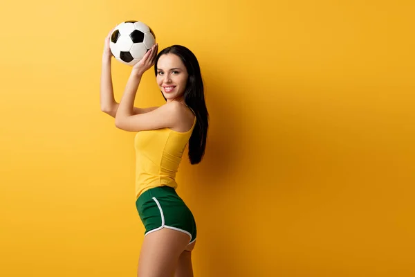 Sorridente fã de futebol feminino em shorts segurando bola no amarelo — Fotografia de Stock