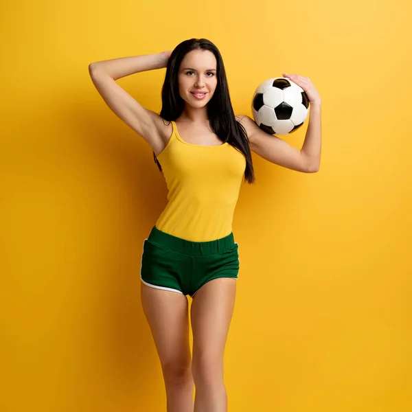 Happy female football fan in shorts holding ball on yellow — Stock Photo