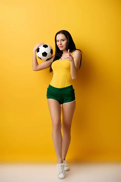 Bela fã de futebol feminino com bola piscando e apontando para você no amarelo — Fotografia de Stock