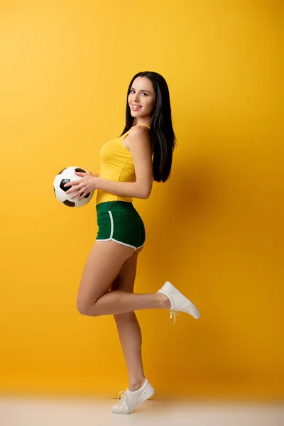 Веселая футбольная фанатка в шортах держит мяч на желтом — стоковое фото