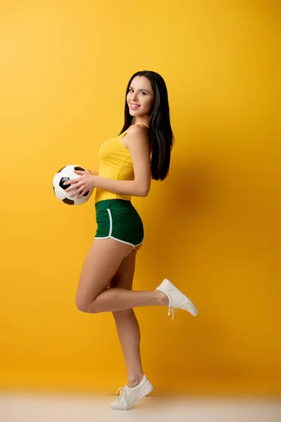 Torcedor de futebol feminino alegre em shorts segurando bola no amarelo — Fotografia de Stock
