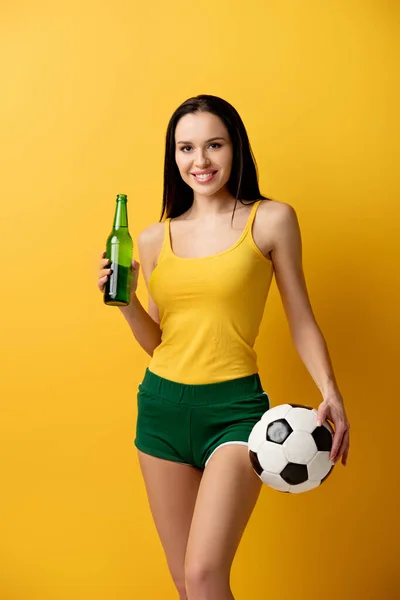 Ventilador de fútbol femenino sonriente sosteniendo bola y botella de cerveza en amarillo - foto de stock