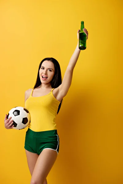 Animado fã de futebol feminino segurando bola e garrafa de cerveja no amarelo — Fotografia de Stock