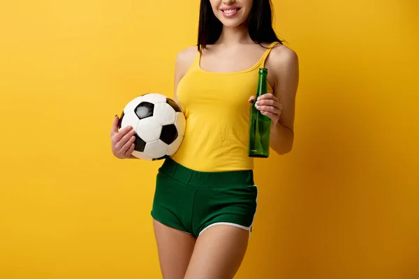 Cropped view of female football fan holding ball and bottle of beer on yellow — Stock Photo