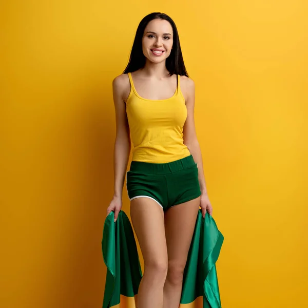Smiling attractive football fan in shorts holding brazilian flag on yellow — Stock Photo