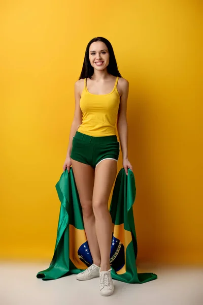 Smiling female football fan in shorts holding brazilian flag on yellow — Stock Photo